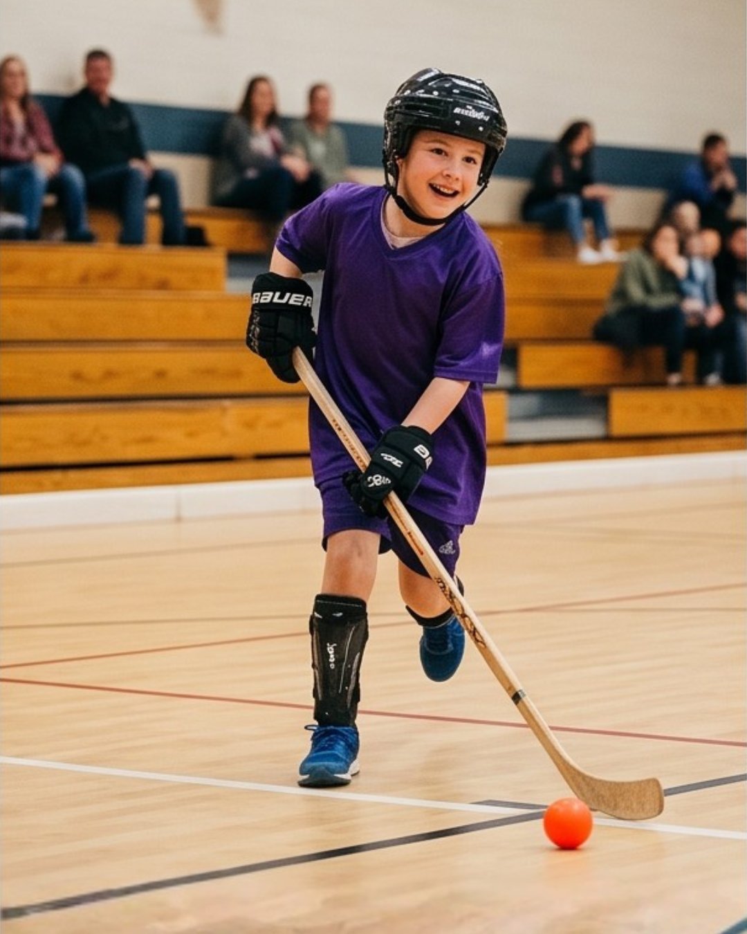 Young hockey player in purple jersey and helmet dribbling orange ball in gymnasium with spectators in bleachers